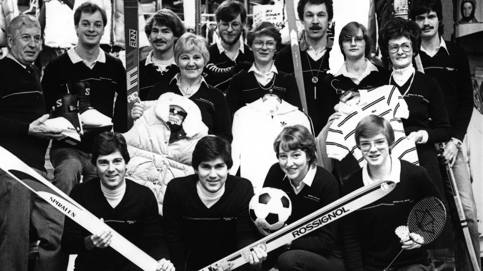A group of people pose together, holding sports equipment like skis, a soccer ball, and a racket, all wearing similar dark shirts.