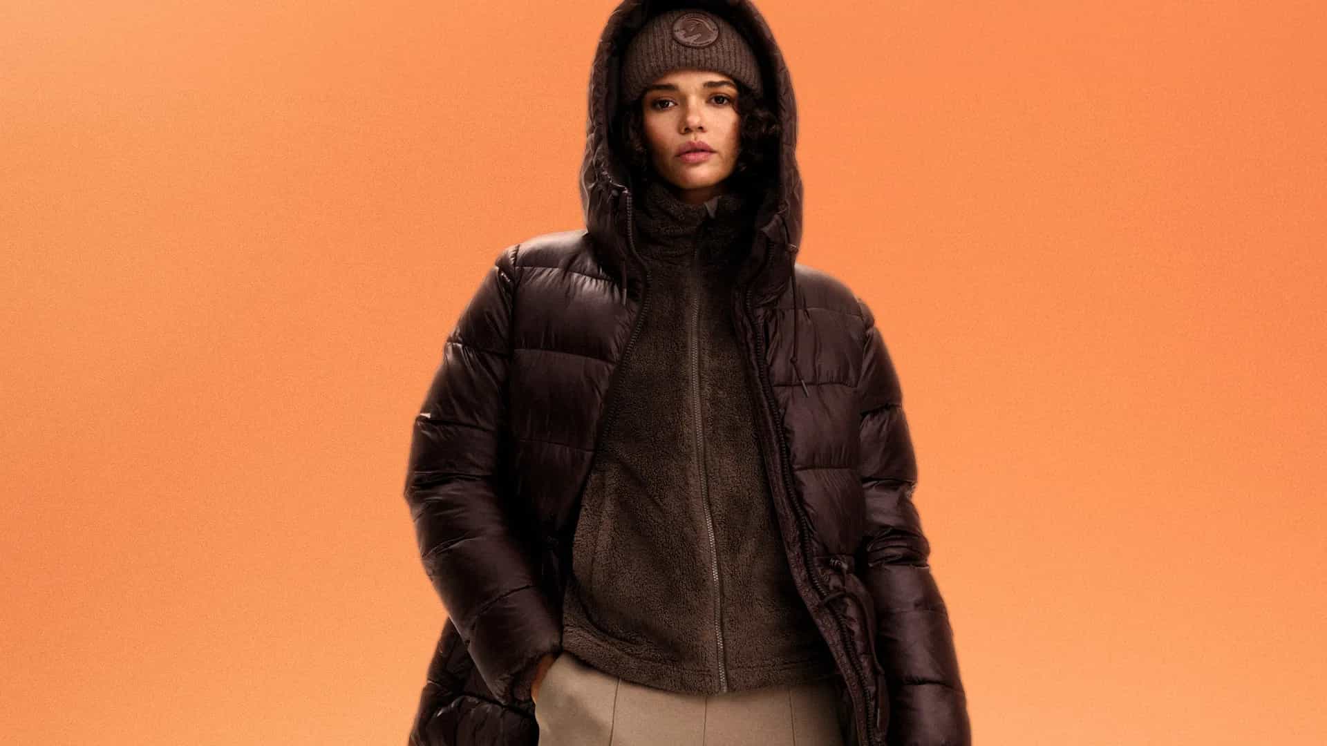 Person in a dark brown puffer jacket and matching hat stands against an orange background.