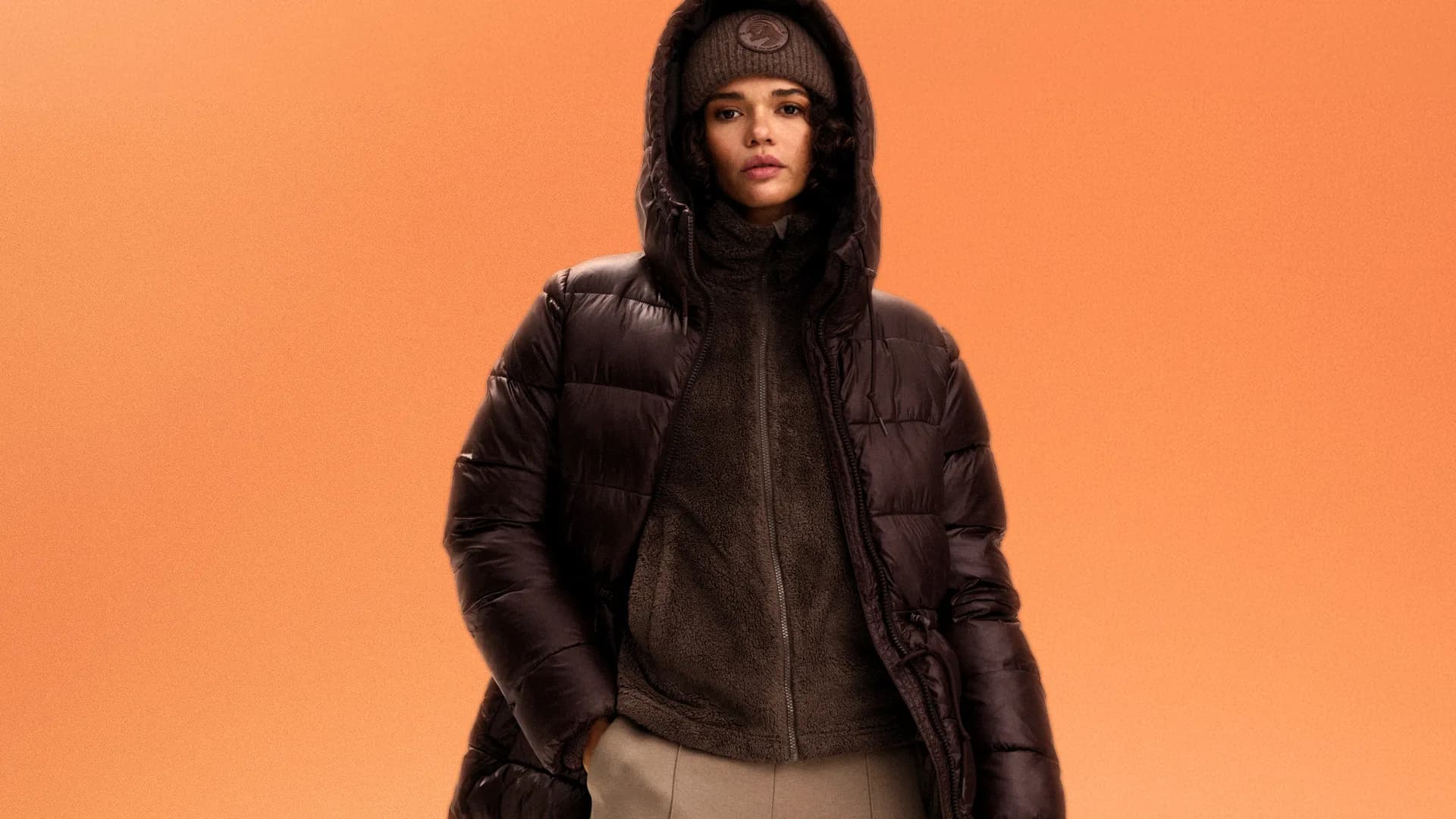 Person in a dark brown puffer jacket and matching hat stands against an orange background.
