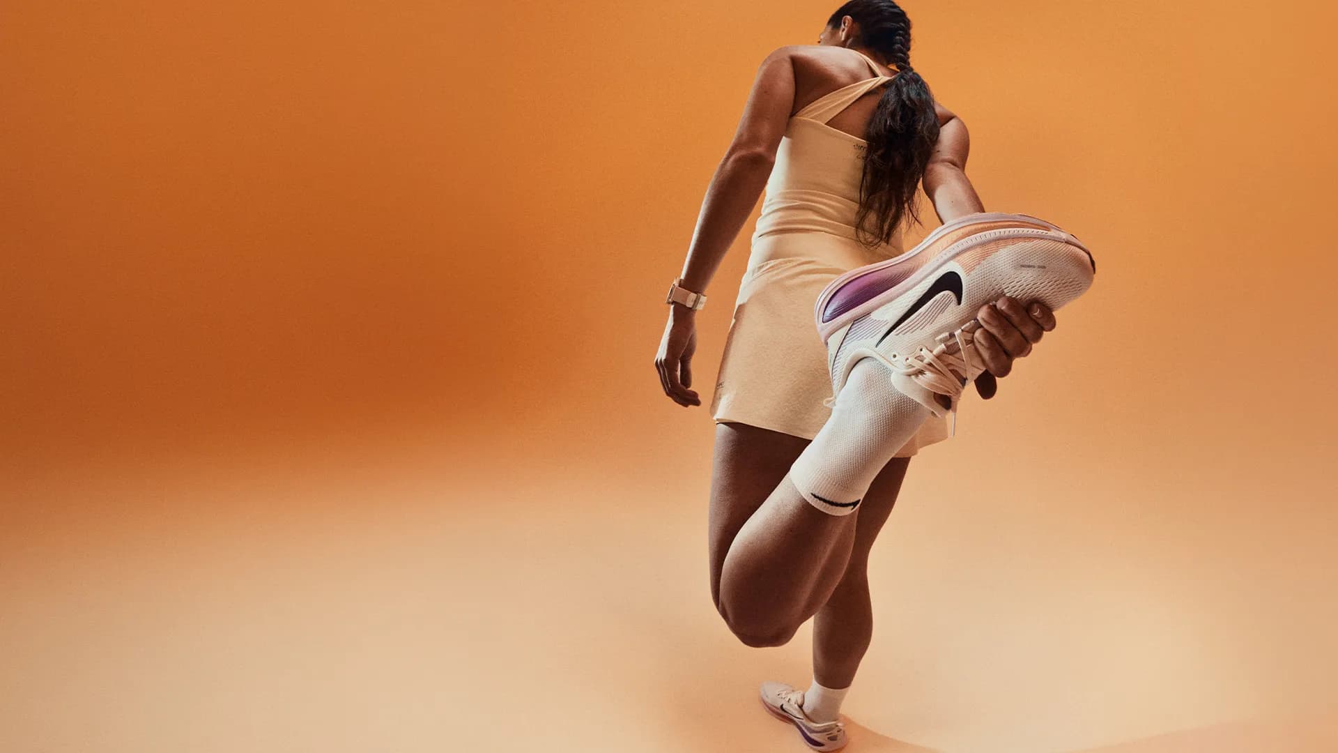 Woman in a sporty outfit stretching her leg, holding a sneaker against an orange background.