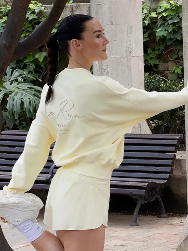 Woman in a light yellow outfit, standing outdoors with one leg raised, near a bench and greenery.